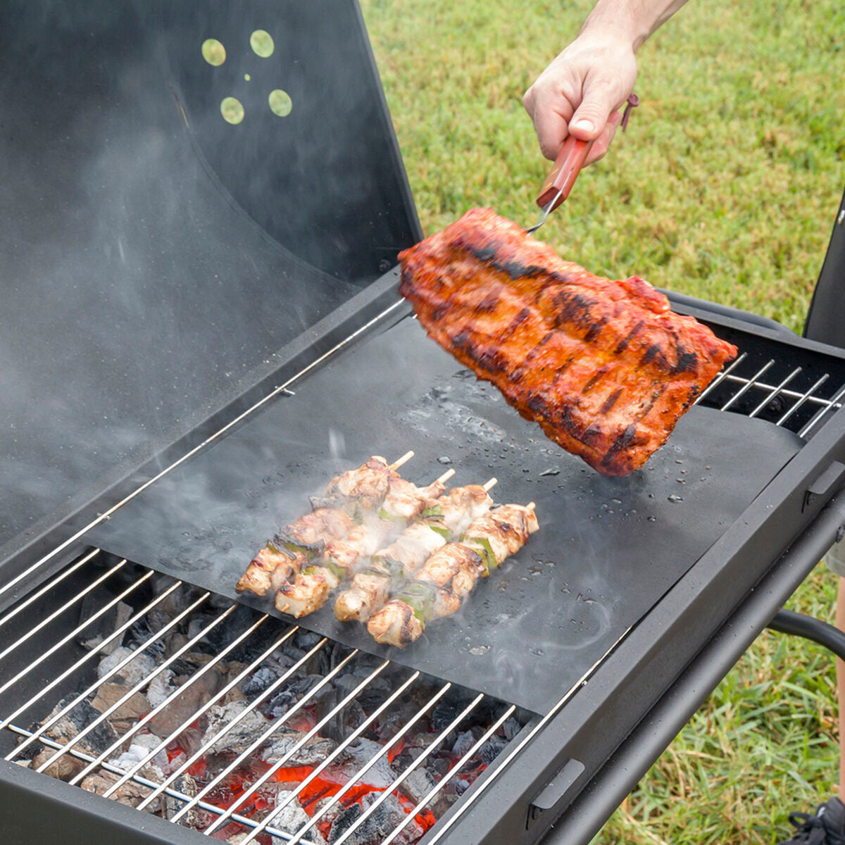 Zwei schwarze InnovaGoods Grillmatten auf einem Grillrost mit Gemüse und kleinen Lebensmitteln