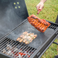 Zwei schwarze InnovaGoods Grillmatten auf einem Grillrost mit Gemüse und kleinen Lebensmitteln